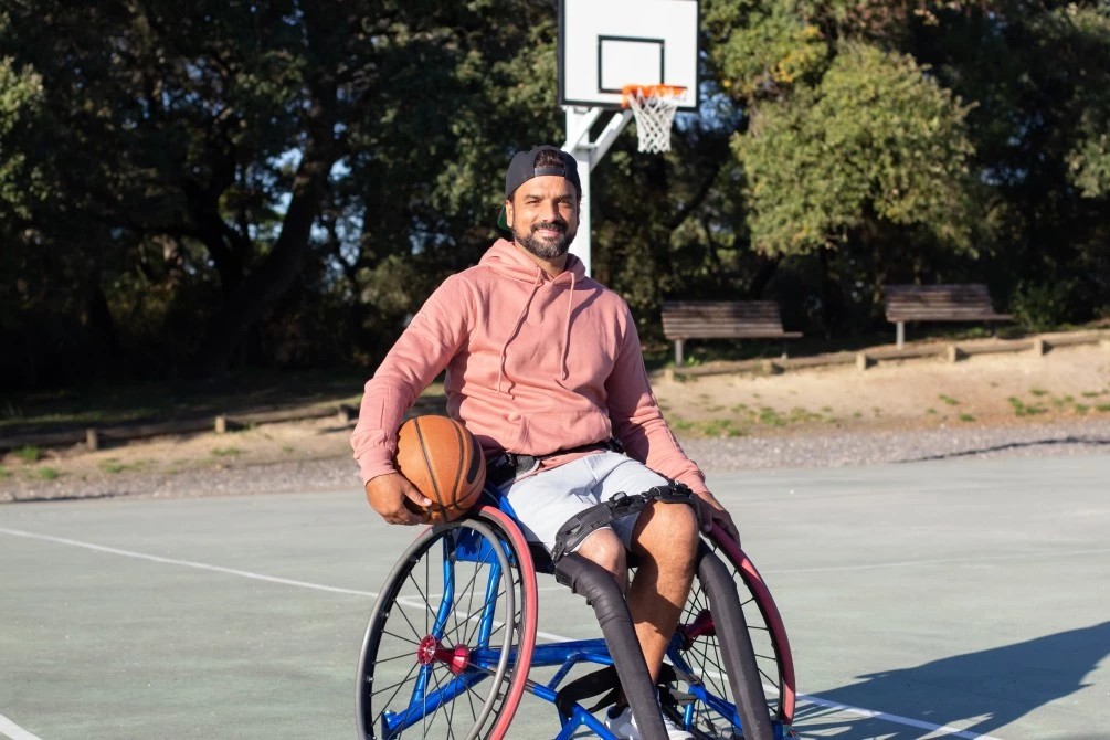 Disabled man in wheelchair on basketball court wearing a cap, holding a basketball, and smiling at camera